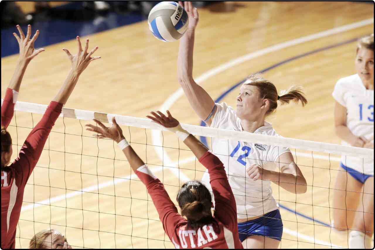 A group of girls jumping to spike a volleyball over the net during a lively match.